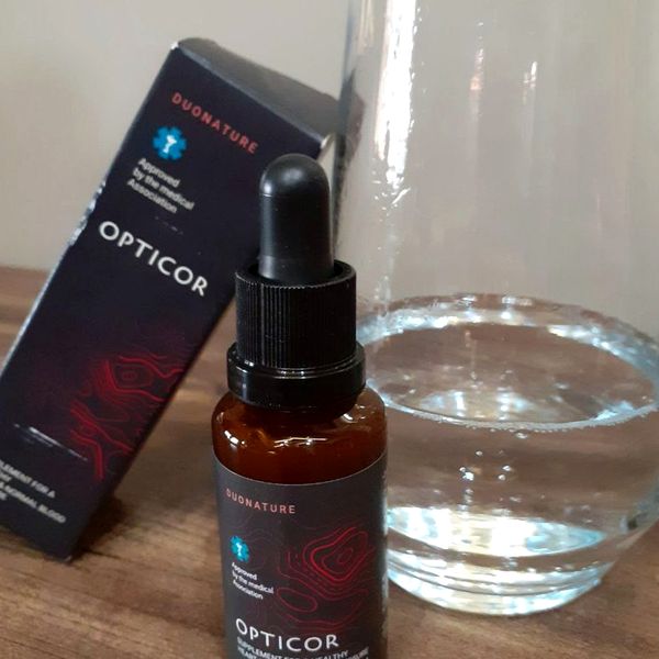 Flacone ambrato di Opticor con contagocce accanto alla scatola e a un bicchiere d'acqua